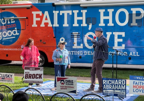 A photo of Doug Pagitt standing in front of the Faith, Hope, & Love bus, speaking.